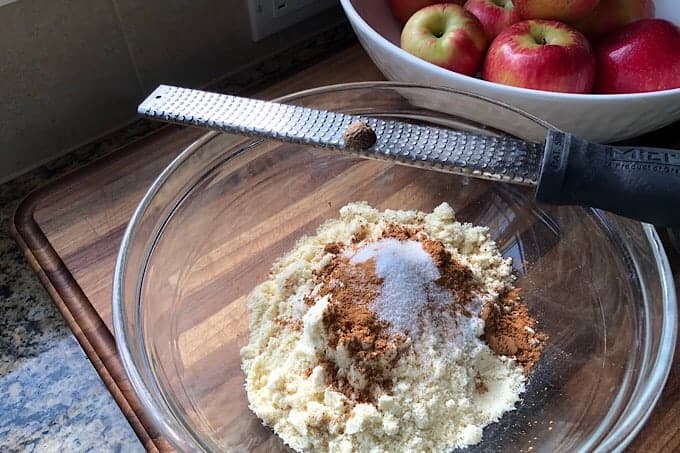 Dry ingredients, unmixed in a clear glass bowl with micro plane and whole nutmeg balanced on side. White bowl of Honeycrisp apples in upper right corner of image.