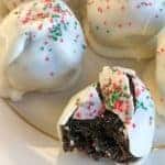 White chocolate balls with red and green sugar sprinkles, placed on a gold rimmed off-white plate. One has a bite out of it revealing soft cream cheese and crushed oreo cookie filling.
