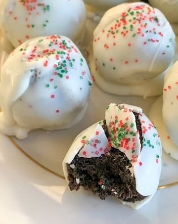White chocolate balls with red and green sugar sprinkles, placed on a gold rimmed off-white plate. One has a bite out of it revealing soft cream cheese and crushed oreo cookie filling.