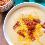A white bowl of delicious Cheesy Potato Soup sprinkled with salty bacon bits and cheddar cheese. Little bowl of bacon bits sits next to soup on an evergreen colored linen napkin.