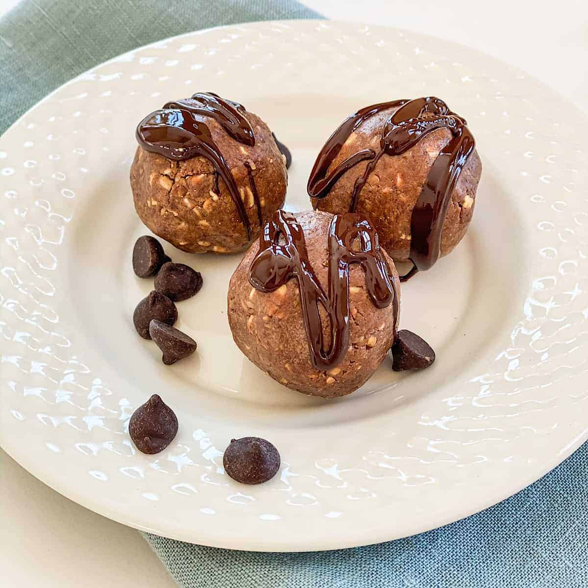 Image of bite sized Chocolate Peanut Butter Protein balls drizzled with chocolate with chocolate chips sprinkled on white plate on light blue linen napkin.