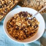 Fresh and delicious homemade granola made with oats, raisins, and honey pictured in a white bowl with spoon placed on blue napkin