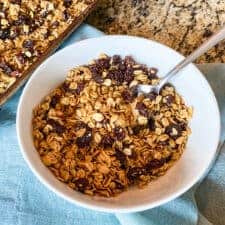 Fresh and delicious homemade granola made with oats, raisins, and honey pictured in a white bowl with spoon placed on blue napkin