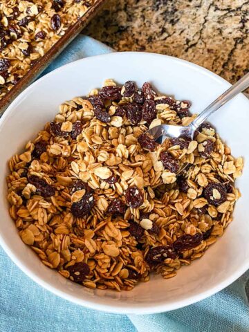 Fresh and delicious homemade granola made with oats, raisins, and honey pictured in a white bowl with spoon placed on blue napkin
