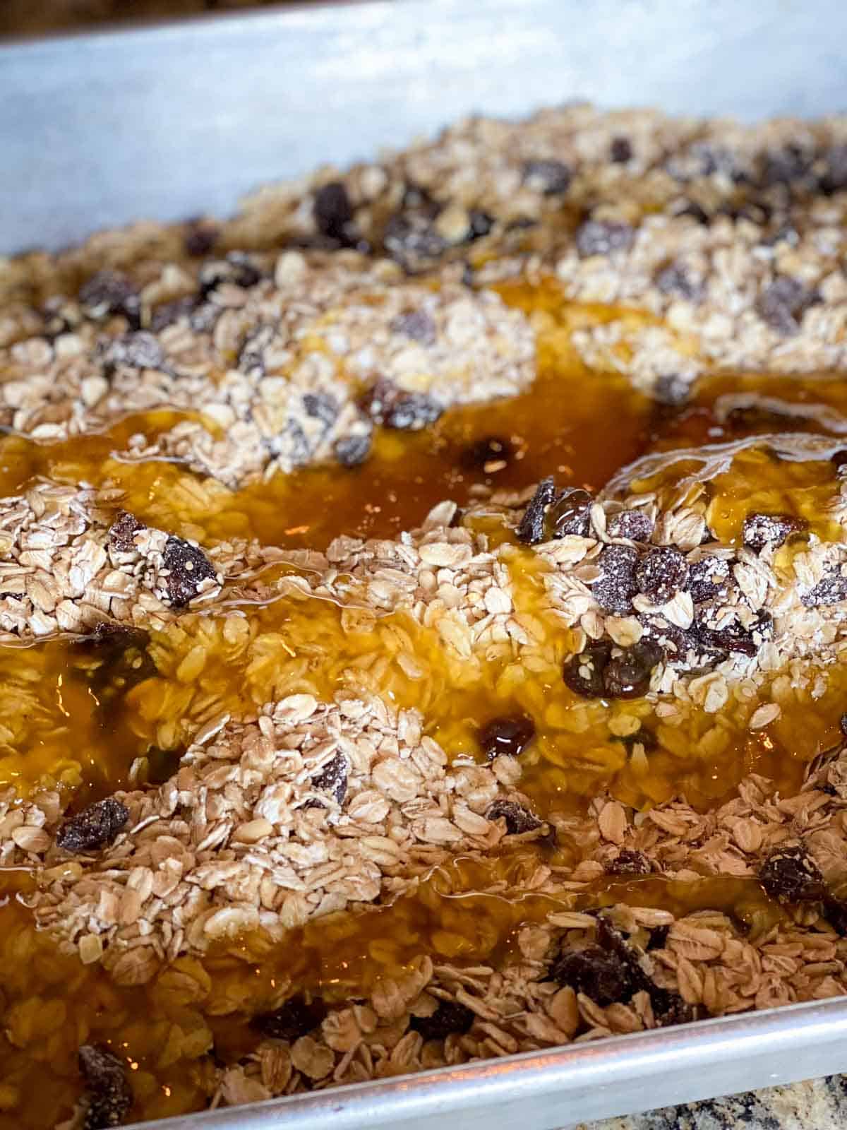 Unbaked honey granola with rolled oats, raisins, and sesame seeds, drizzled with honey and olive oil before mixing.