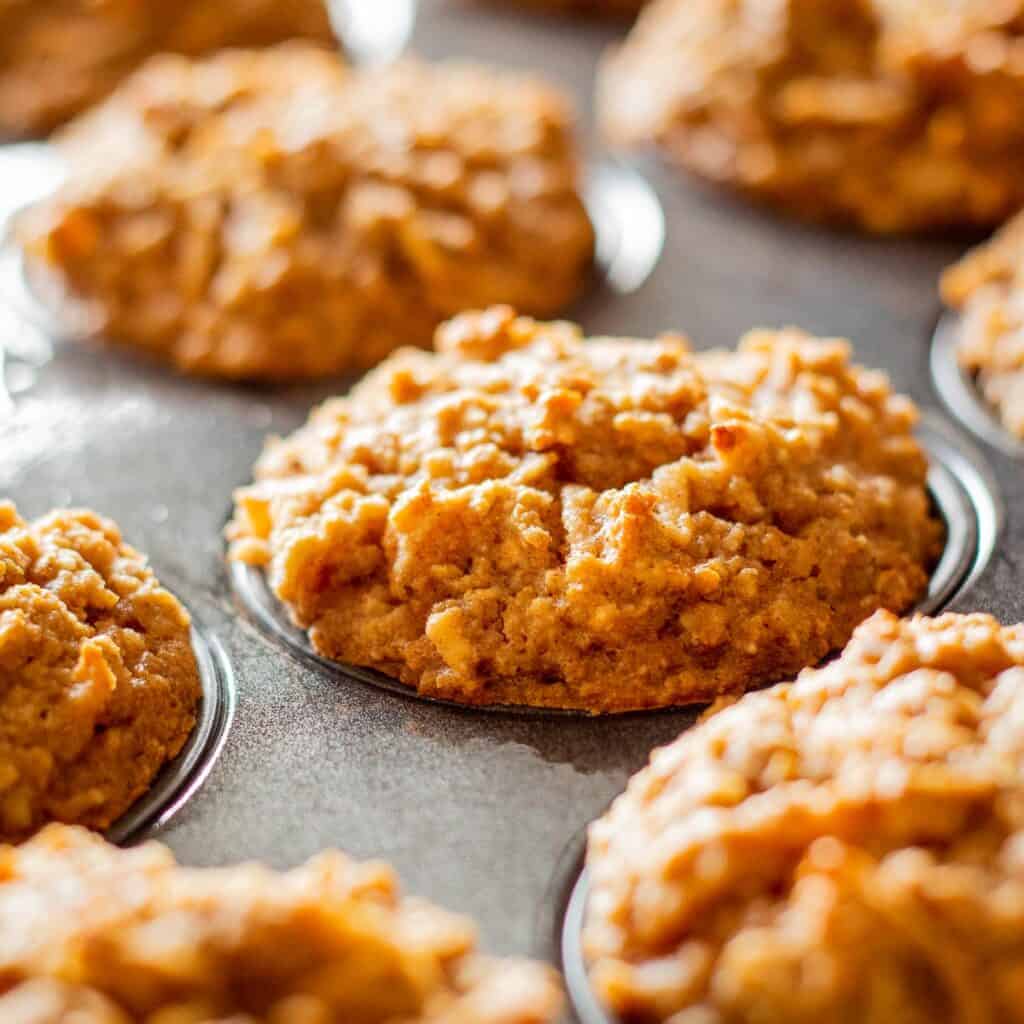 One fresh apple muffin placed on a white plate with an apple. Muffin tin filled with muffins is visible in background.