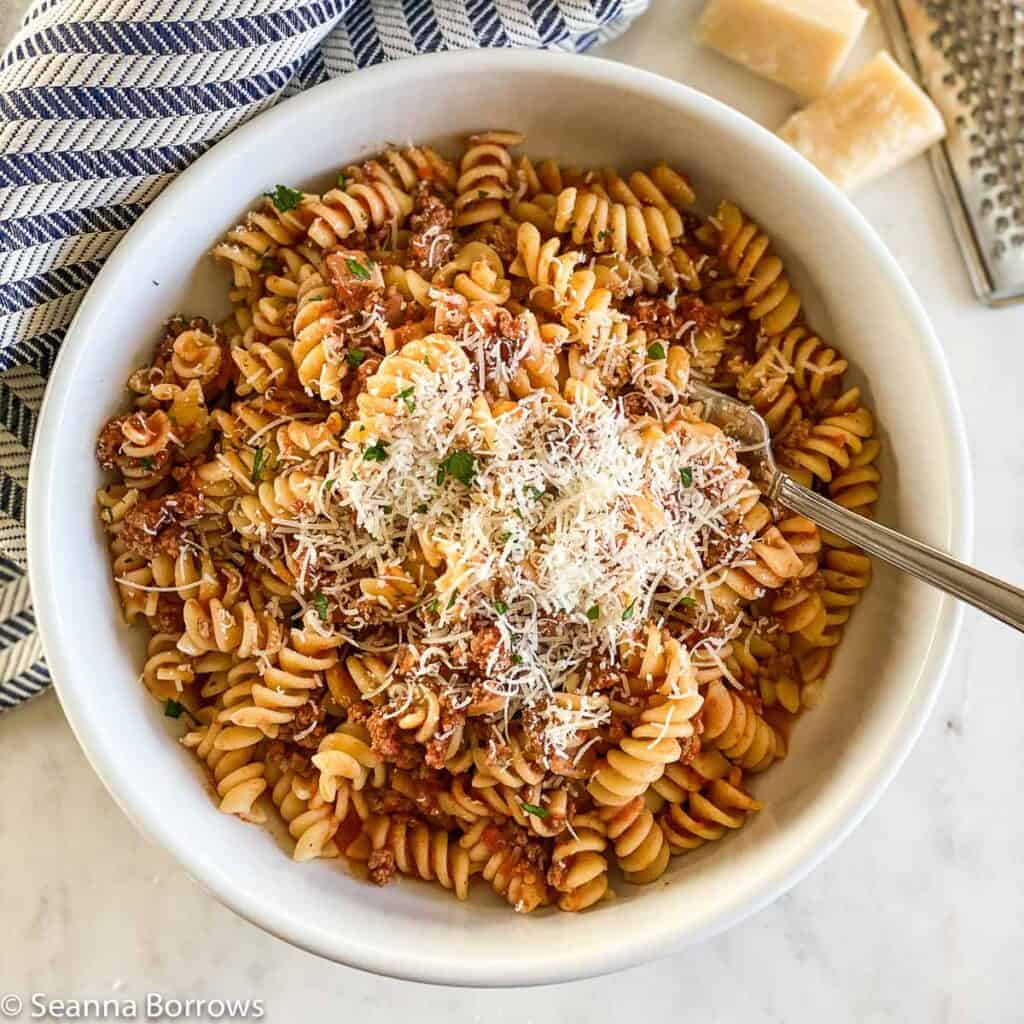 Bison Pasta made with rotini pictured in a white bowl, topped with freshly grated Parmigiano-Reggiano. Fork in bowl on right side. Muted blue and white stripped towel placed on upper left side of image. Three small pieces of Parmigiano-Reggiano pictured in upper right corner next to a small micro-plane grater.