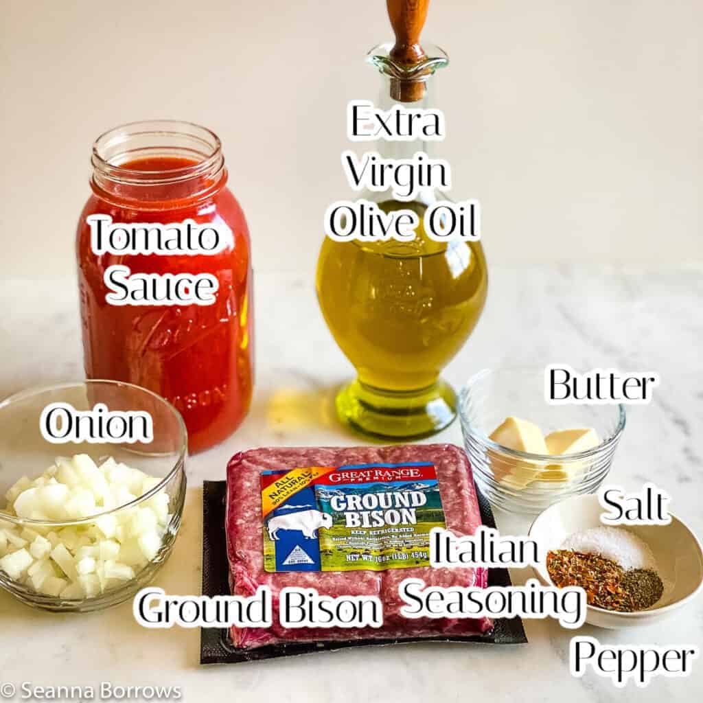 Ingredients for Bison Pasta arranged in circle top clockwise: tall glass container of Olive Oil, small clear dish of 2 tablespoons butter, small off white dish of salt, pepper, and Italian Seasonings, 1 pound package of ground bison, small glass bowl of Parmigiano-Reggiano, 32 ounce glass Ball canning jar of red tomato sauce