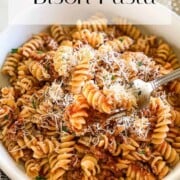 Bison Pasta made with rotini pictured in a white bowl, topped with freshly grated Parmigiano-Reggiano. Fork in bowl on right side. Muted blue and white stripped towel placed on upper left side of image. Three small pieces of Parmigiano-Reggiano pictured in upper right corner next to a small micro-plane grater.