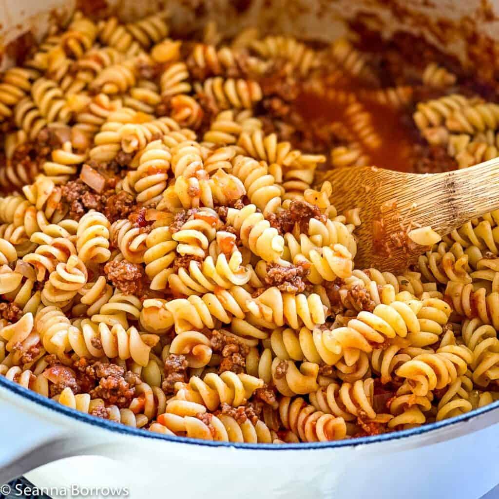Bison Pasta made with rotini pictured in off white Le Creuset dutch oven with wooden spoon gently lifting up some of the yummy pasta.