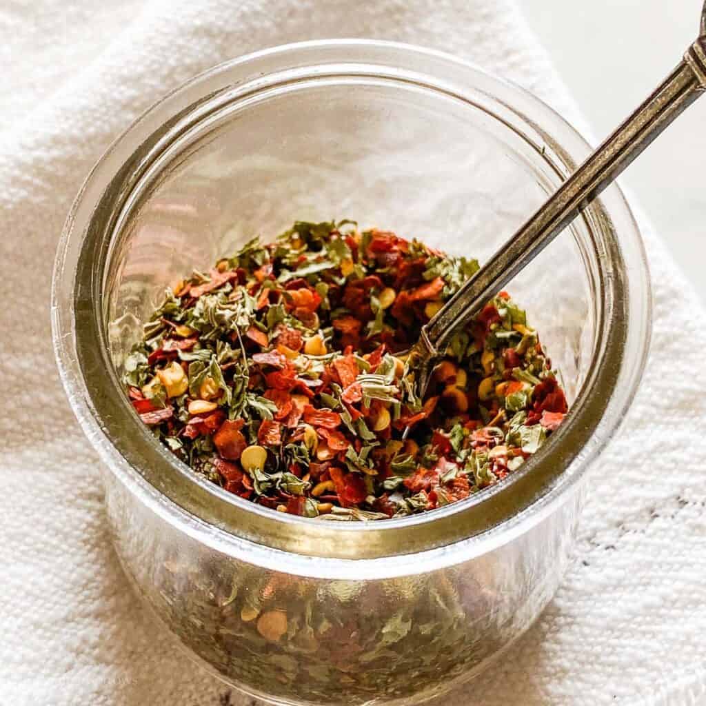 Red and green Italian Seasoning mixture pictured in a clear glass straight sided cup with little antique spoon. Pictured on a cream colored fringed napkin.