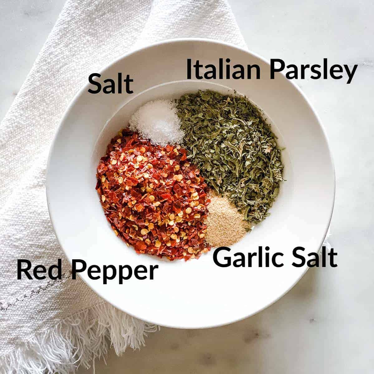 White bowl with spices arranged in a circle and labeled clockwise from top: Italian Parsley, Garlic Salt, Red Pepper, and Salt. Bowl is placed on a off white, fringed napkin on a white marble background.