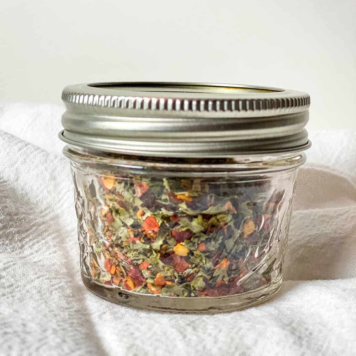 Pretty Red and Green Italian Seasoning stored in 4 oz mason jar with silver screw top lid, placed on white floursack towel against white background.