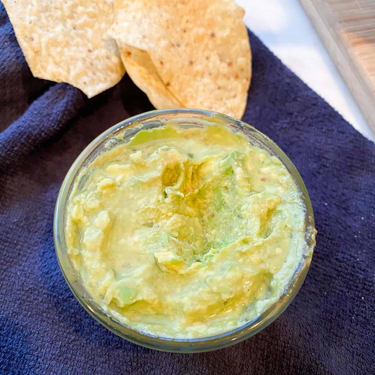 Image of a small bowl of fresh, green, perfectly preserved guacamole using our Make Ahead Secret for Guacamole placed on blue towel with chips on side. Delish!