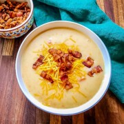 A white bowl of delicious Cheesy Potato Soup sprinkled with salty bacon bits and cheddar cheese. Little bowl of bacon bits sits next to soup on an evergreen colored linen napkin.