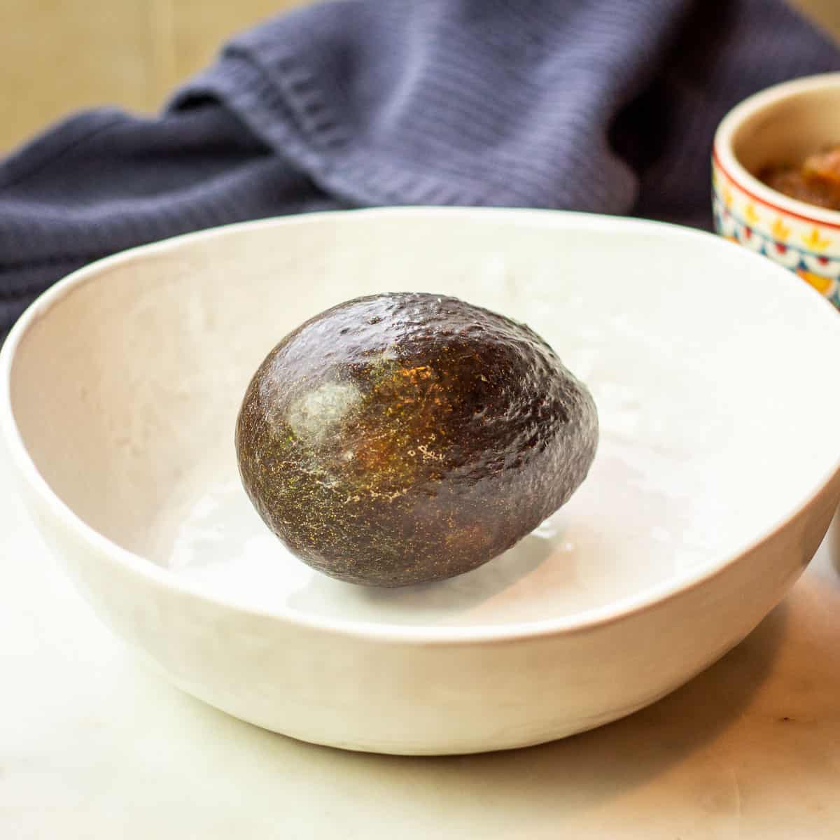 Image of a perfectly ripe avocado pictured in a white bowl with blue kitchen towel nearby.