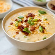 Image of a white bowl of Cheesy Potato Soup with sprinkles of bacon bits, shredded cheese, and green onion.