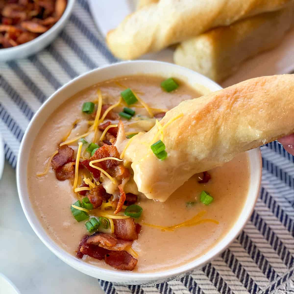 Image of a breadstick dipping into a bowl of The Best Potato Soup topped with cheddar cheese, bacon, and green onion.
