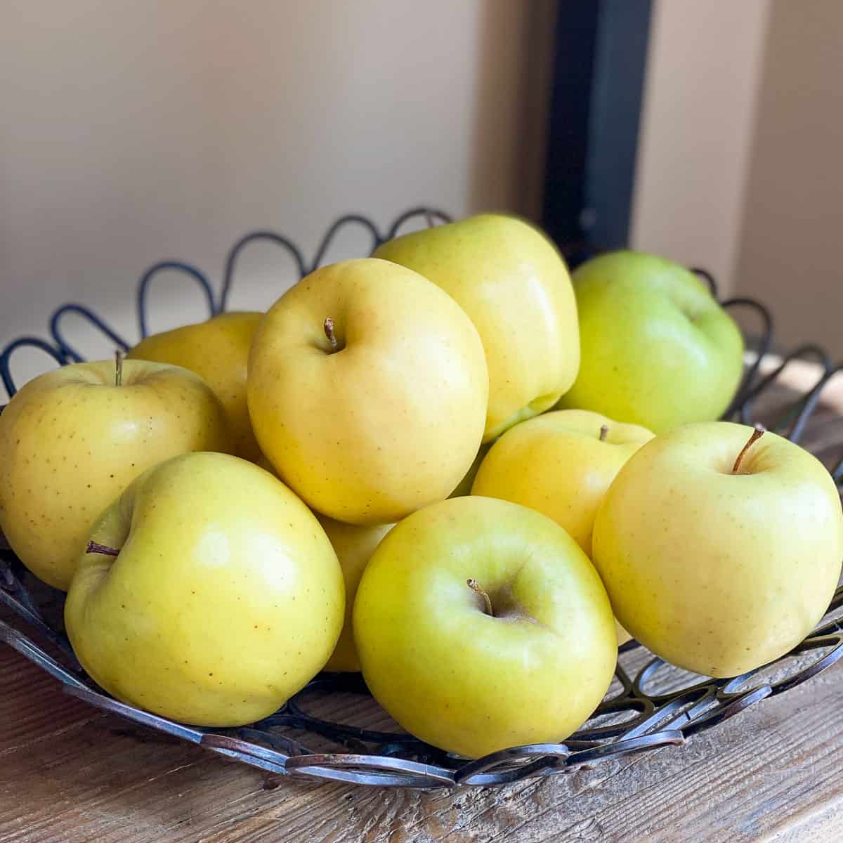 A decorative wire bowl of Golden Delicious Apples on a wooden shelf.