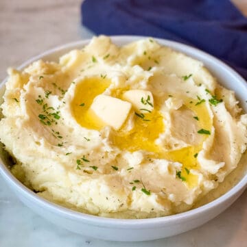 Make-Ahead Mashed Potatoes shown in white serving bowl with melting tabs of butter and fresh chopped Italian Parsley garnish.
