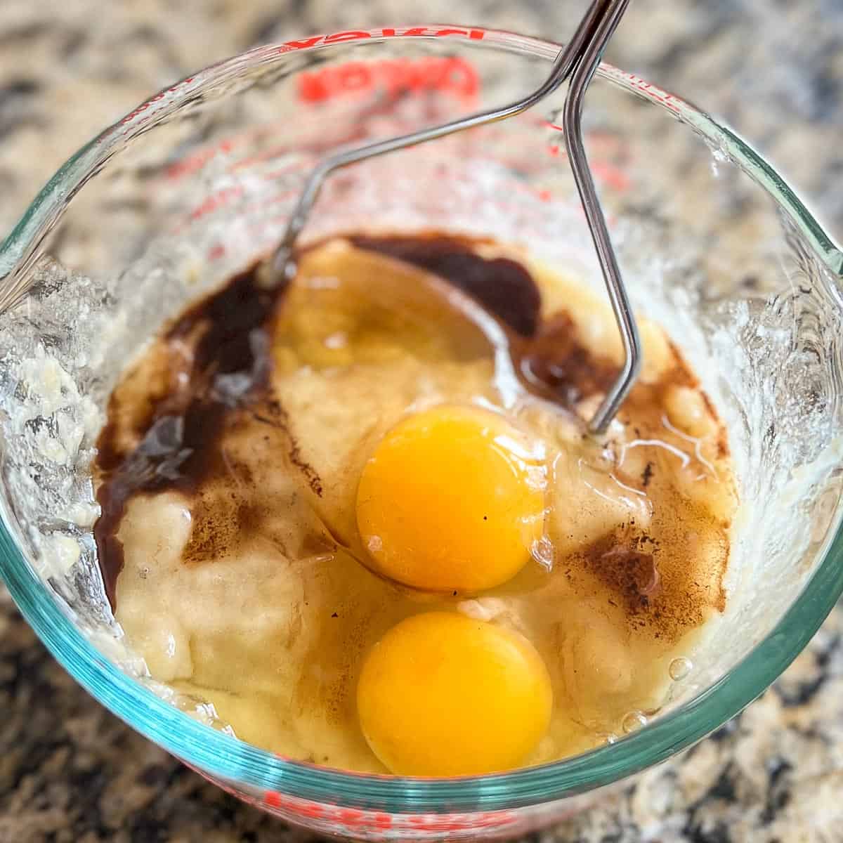 Mashed bananas with two cracked eggs and vanilla extract in a glass measuring cup with a masher.