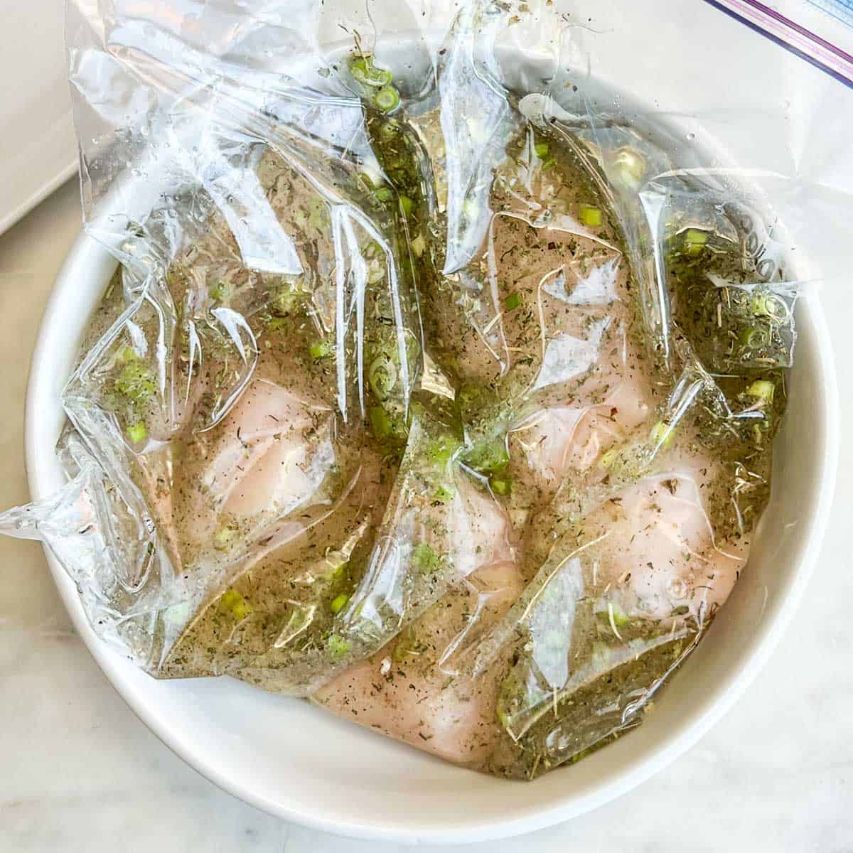 Chicken marinating in a ziploc bag set in a white bowl.