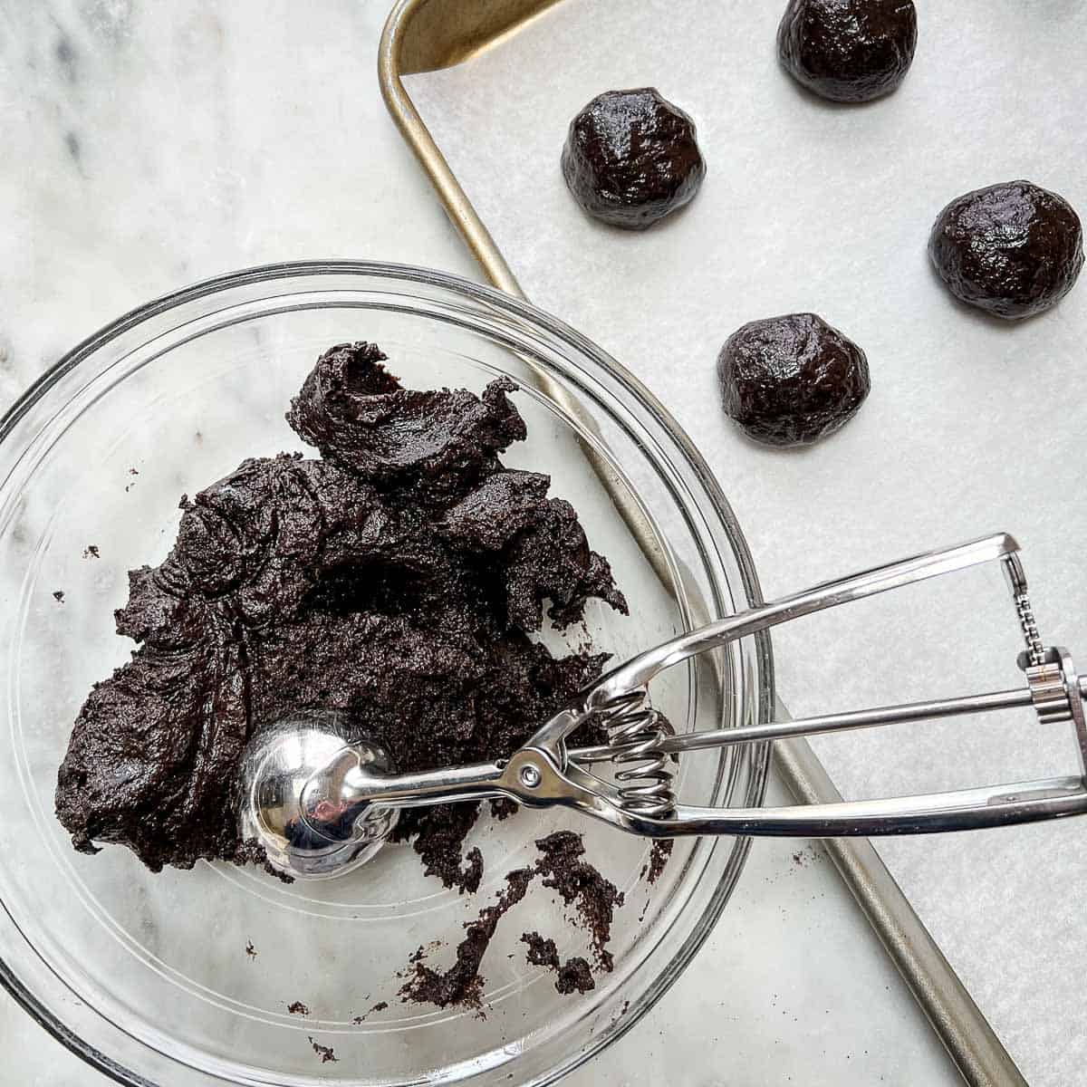 Bowl of Oreo Truffle dough with small scoop inside clear glass bowl next to sheet of small oreo balls.