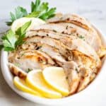 A small white platter of sliced oven roasted turkey breast garnished with lemons and fresh parsley.