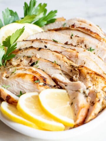 A small white platter of sliced oven roasted turkey breast garnished with lemons and fresh parsley.