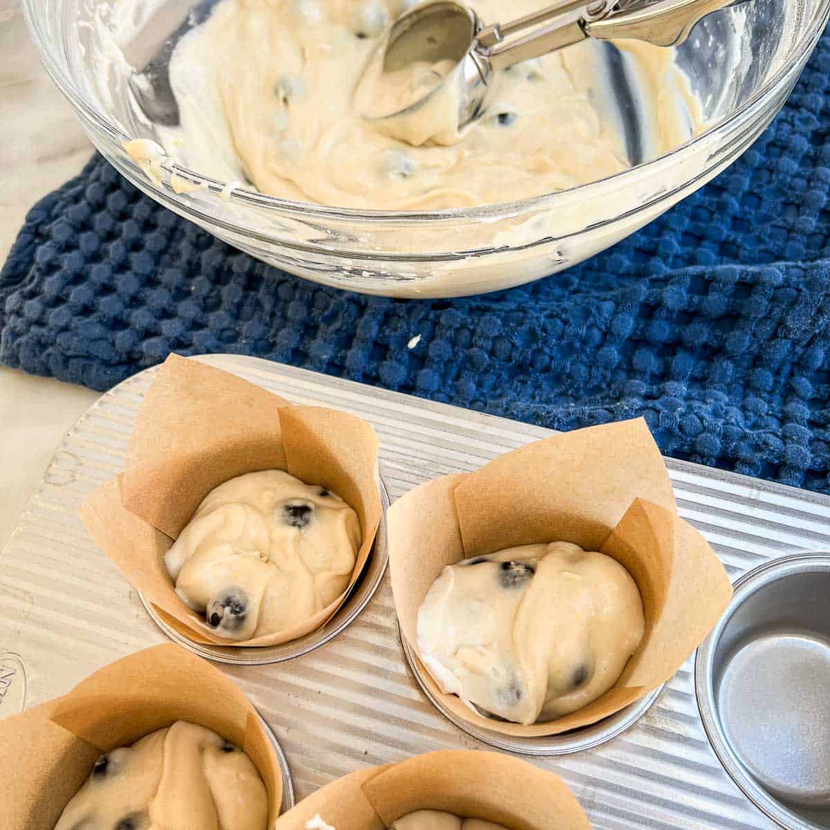 A muffin pan being filled with blueberry muffin batter with a bowlful of batter and a cookie scoop.