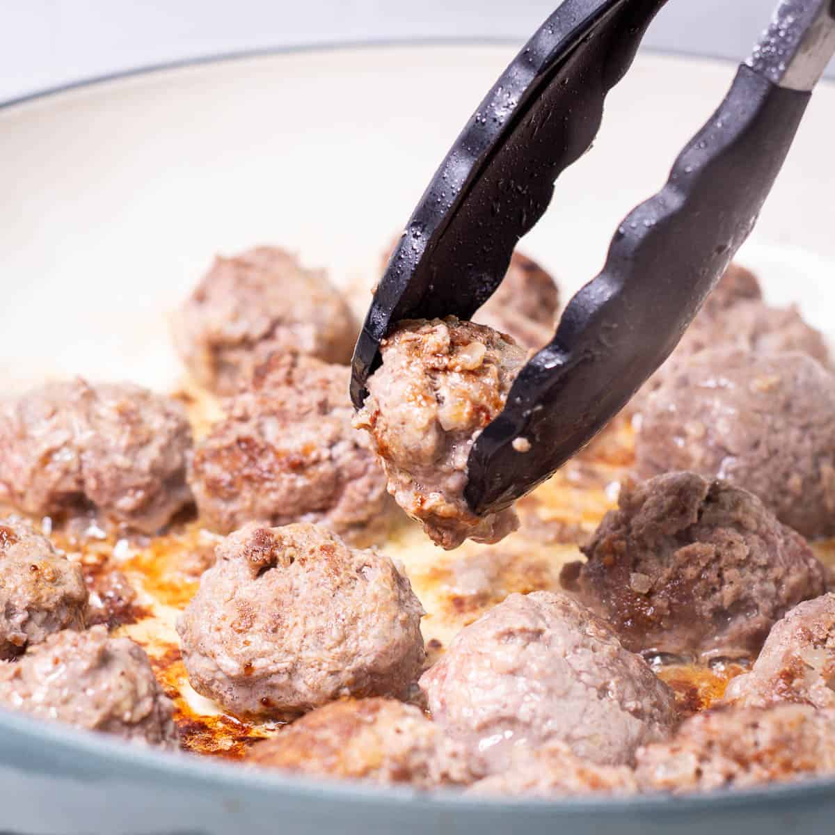 Bison meatballs simmering in a pan with one meatball being raised with kitchen tongs.