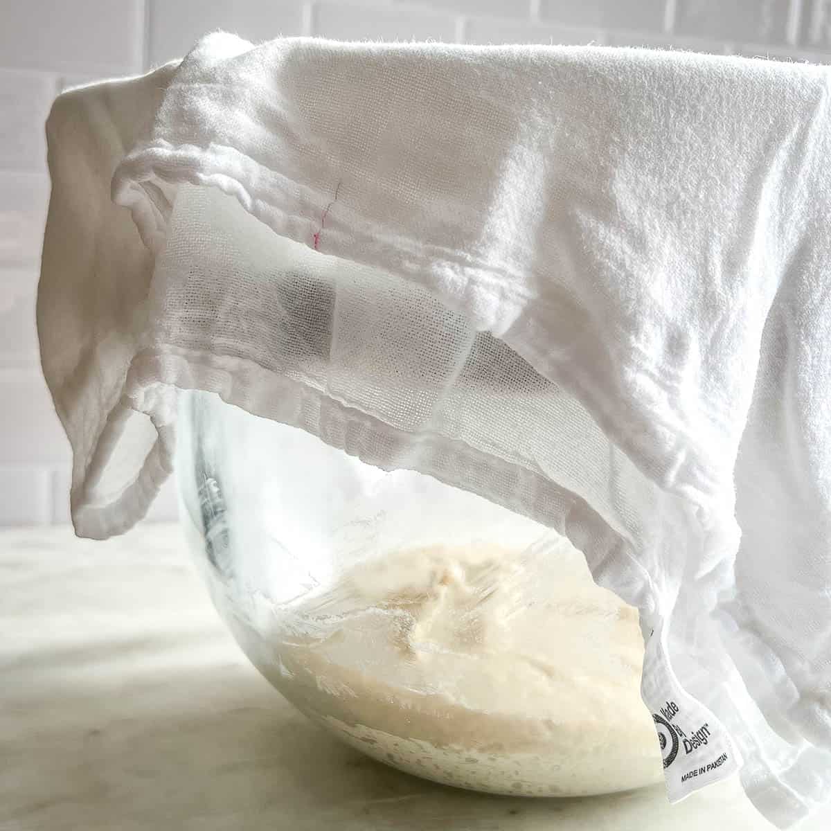 Cinnamon Roll dough in a clear glass stand mixing bowl with a white towel covering it for the first rise.