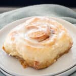 A freshly baked cinnamon roll with icing on top pictured on a white plate with light blue napkin.