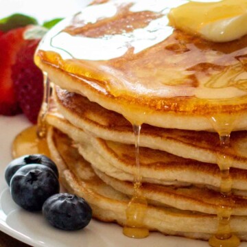 A stack of five buttermilk pancakes with butter on top and maple syrup dripping down the stack with berries on the side.