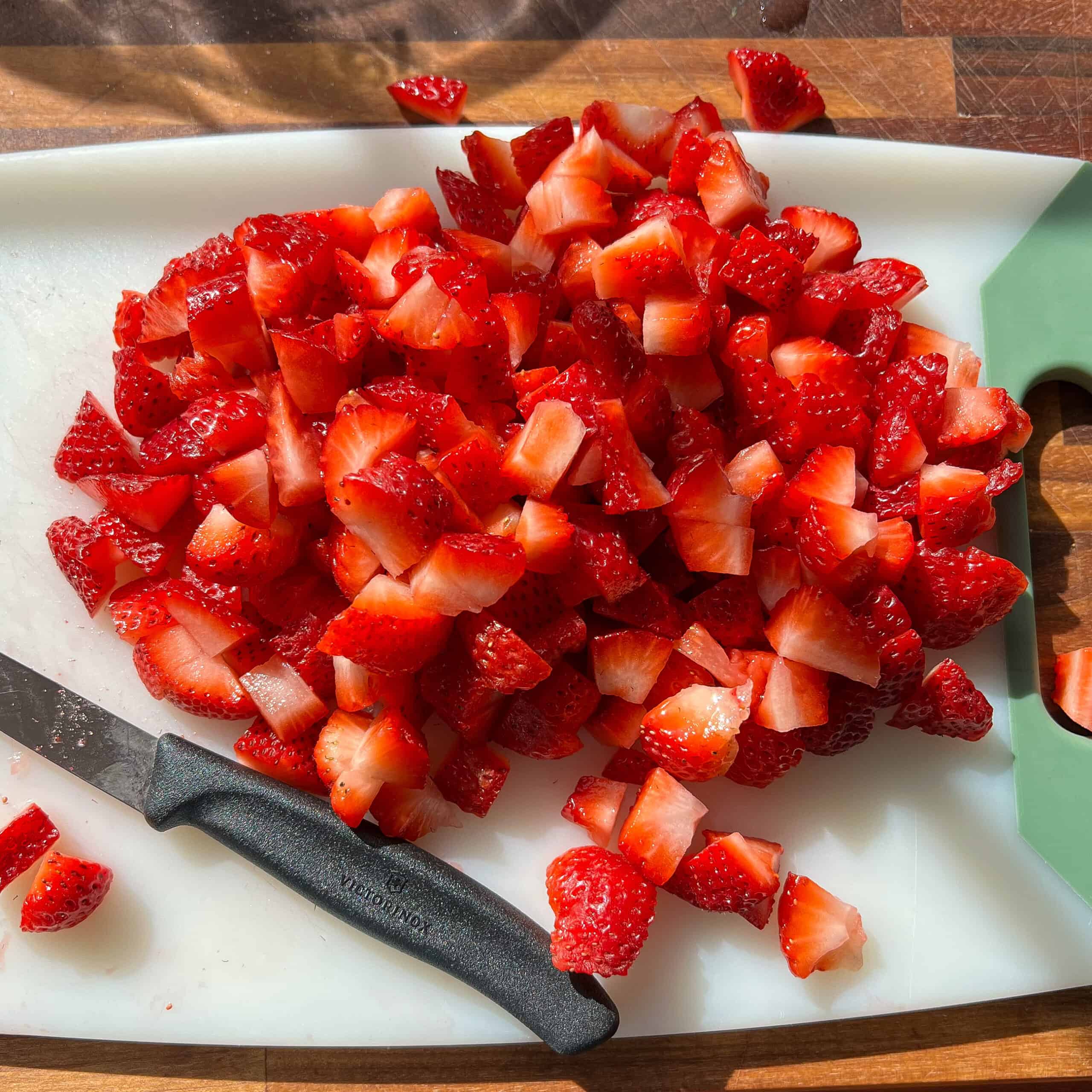 Chopped strawberries on a white cutting board with a pairing knife to the left side.