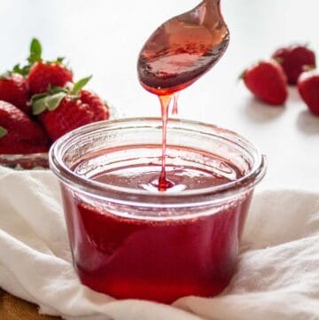 Homemade strawberry syrup dripping from spoon into dish with strawberries in background.