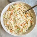 The best coleslaw for pulled pork in a white bowl with a spoon and a light blue towel to the side.