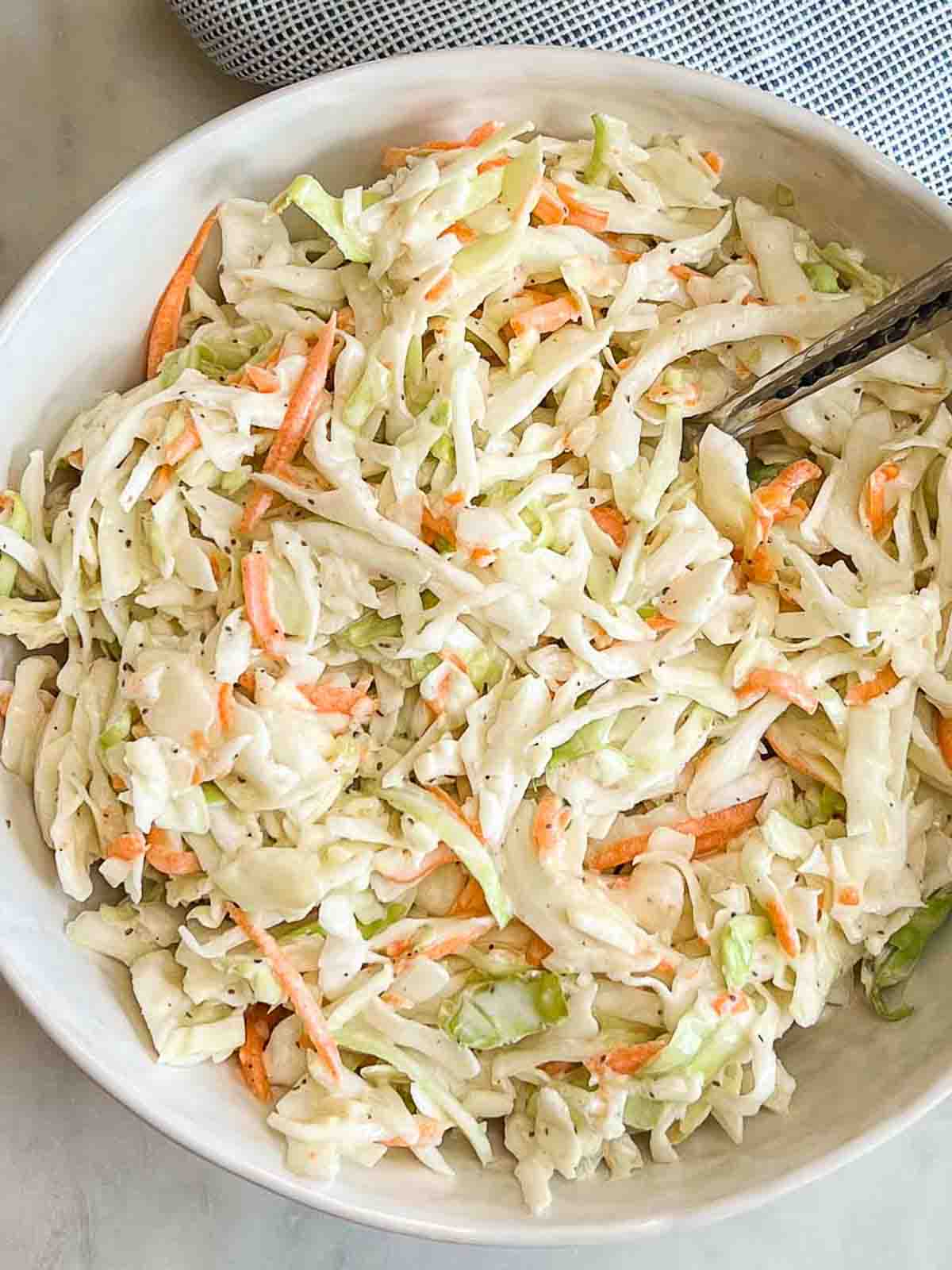 Creamy homemade coleslaw in a white serving bowl with a spoon and light blue towel beside it.