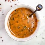 A small, white dipping bowl full of zesty Cowboy Butter Sauce with a little spoon inside bowl and spices scattered around.
