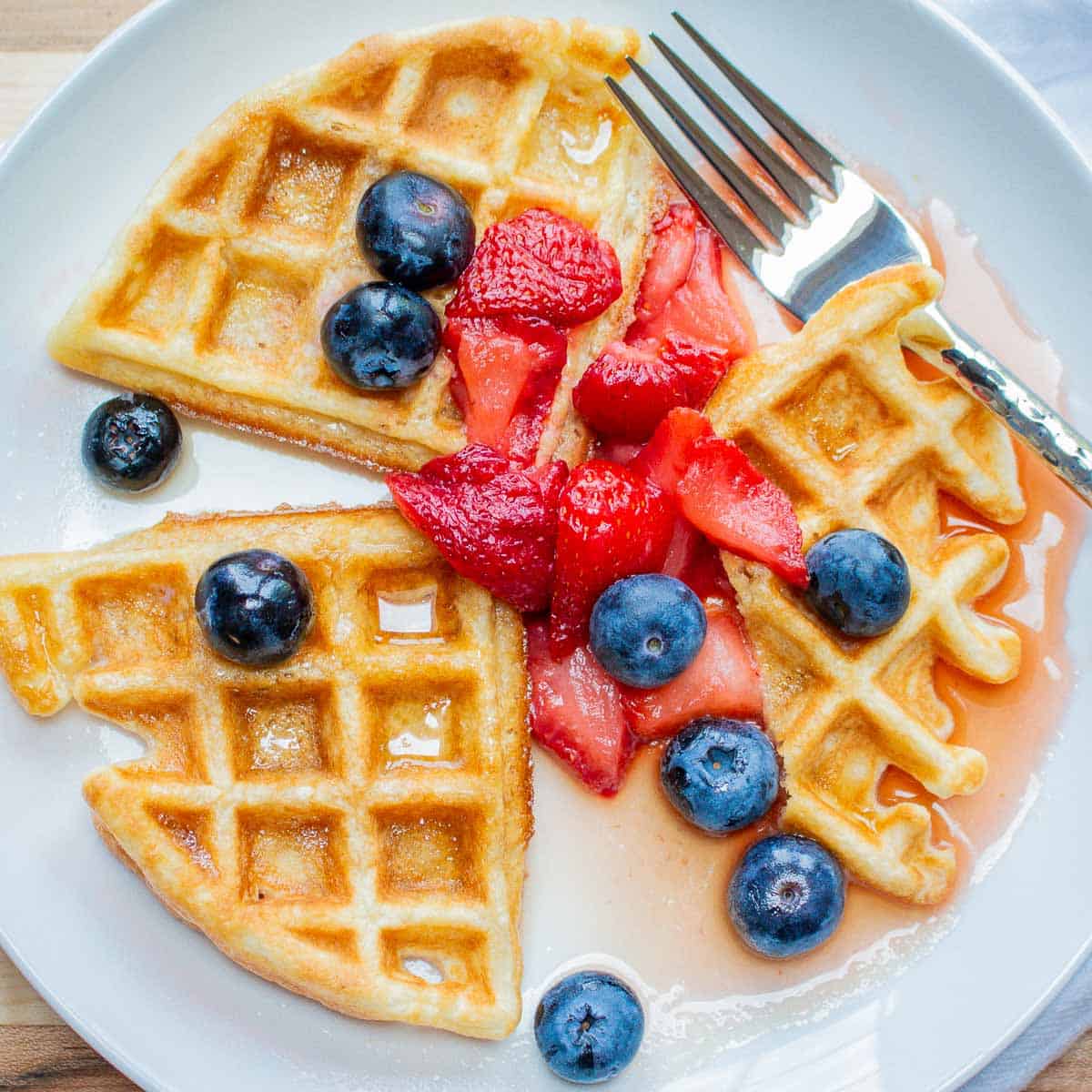 Three pieces of a Homemade Waffle on a white place with strawberries and blueberries and a little maple syrup in the wells of the waffle.