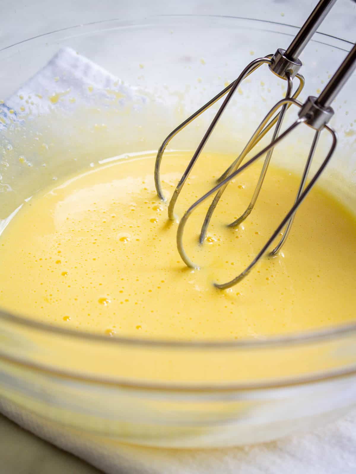 A bowlful of egg yolks and sugar that have been beaten into a pale yellow color with hand mixer whisk attachments shown.