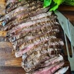 A whole grilled marinated flank steak that has been sliced and fanned out on a cutting board with parsley garnish and a large carving knife and fork on the side.