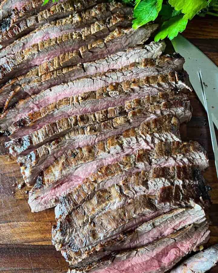 A whole grilled marinated flank steak that has been sliced and fanned out on a cutting board with parsley garnish and a large carving knife and fork on the side.