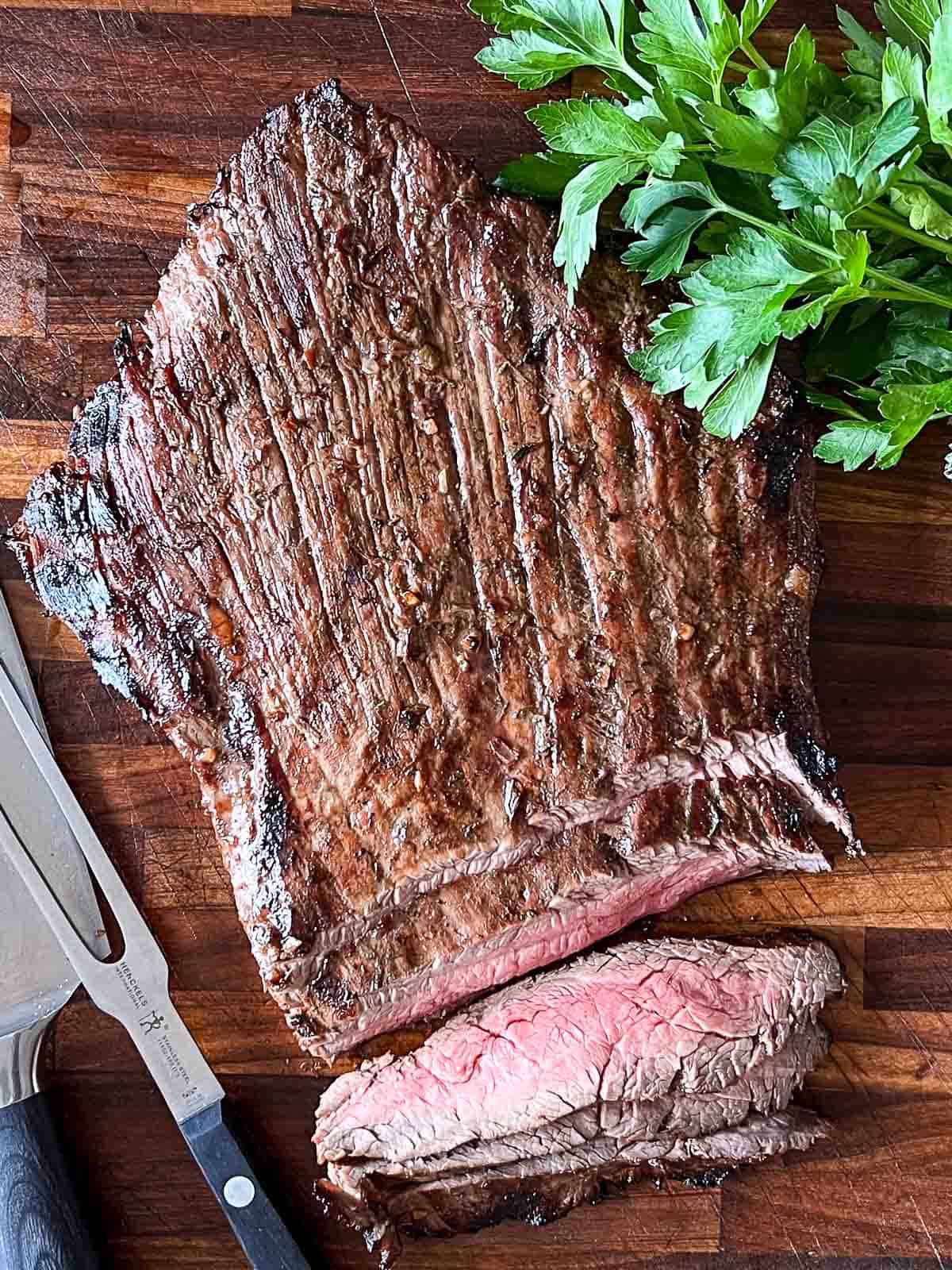 A whole grilled marinated flank steak with a few slices on a cutting board with Italian parsley garnish.