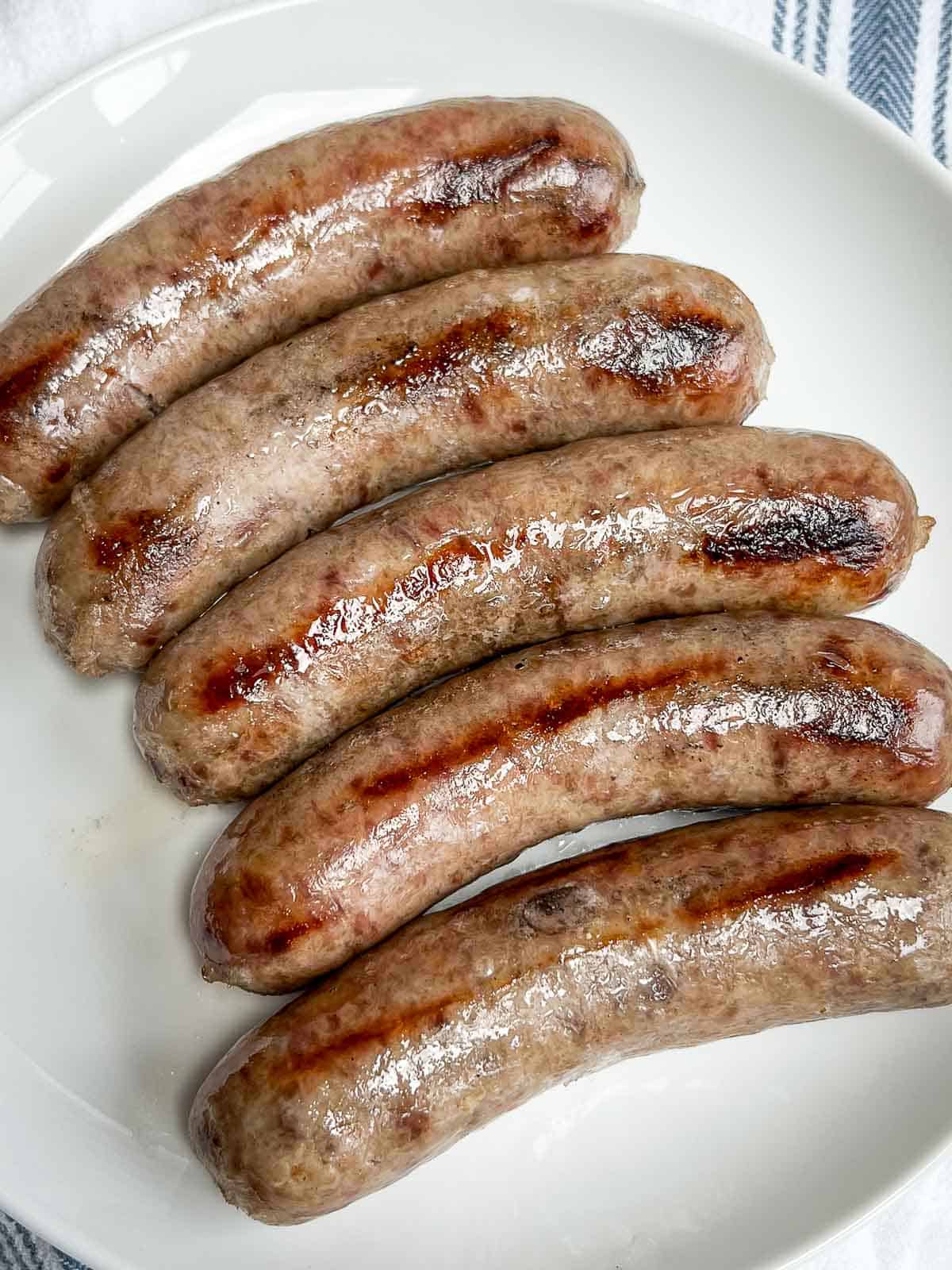 5 grilled beer brats lined up on a white plate.