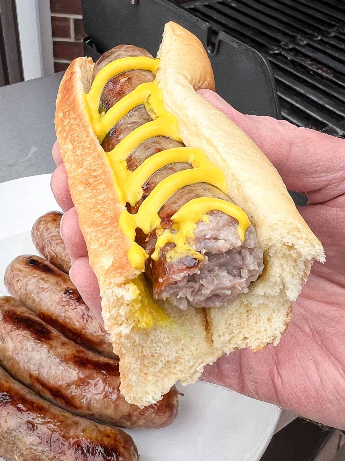 A grilled beer brat with yellow mustard in a bun being held up to the camera.