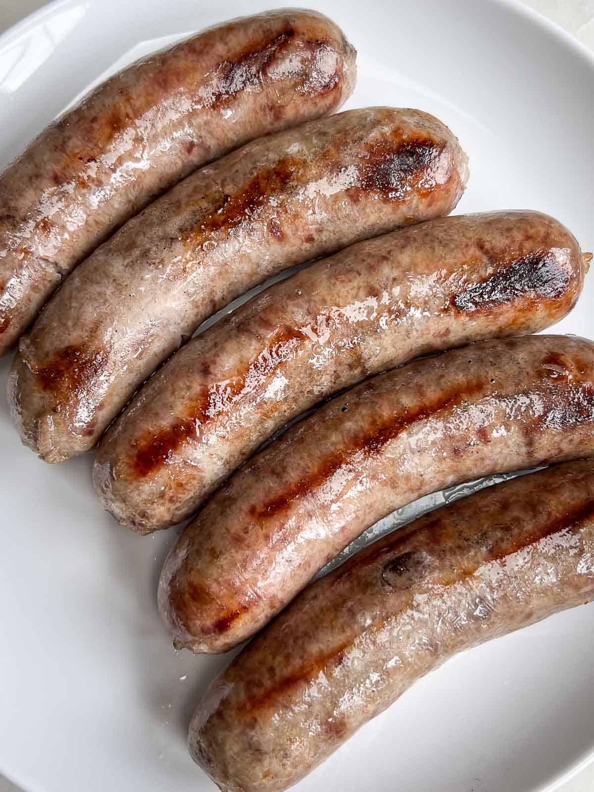 5 grilled beer brats lined up on a white plate.