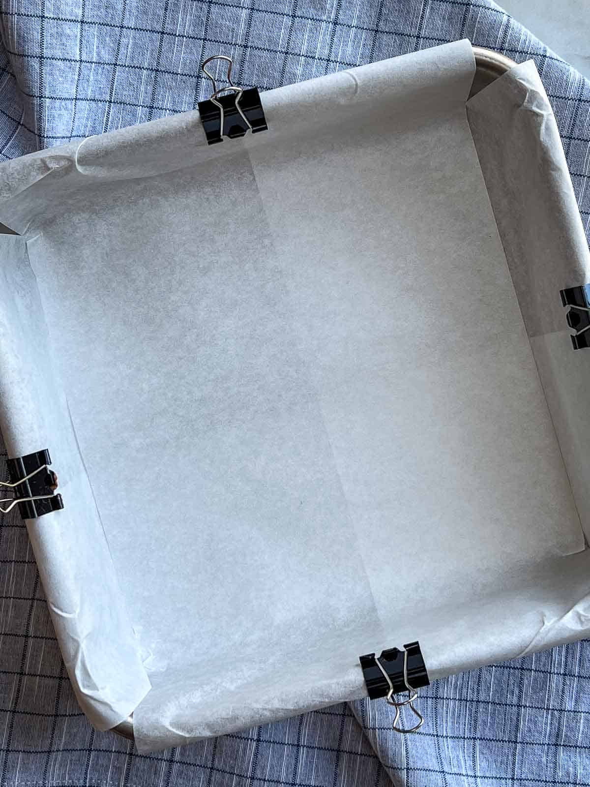 Square baking pan that has been lined with parchment paper held in place by a binder clip on the each side.