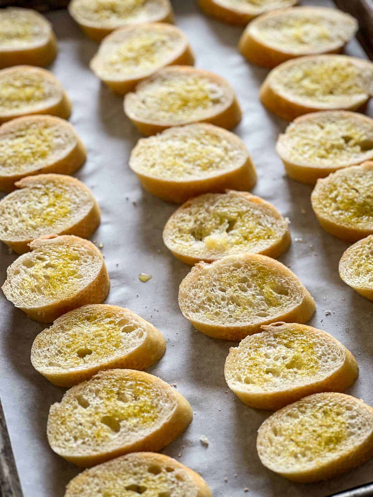 Thin slices of baguette topped with olive oil on a sheet pan ready for the oven.