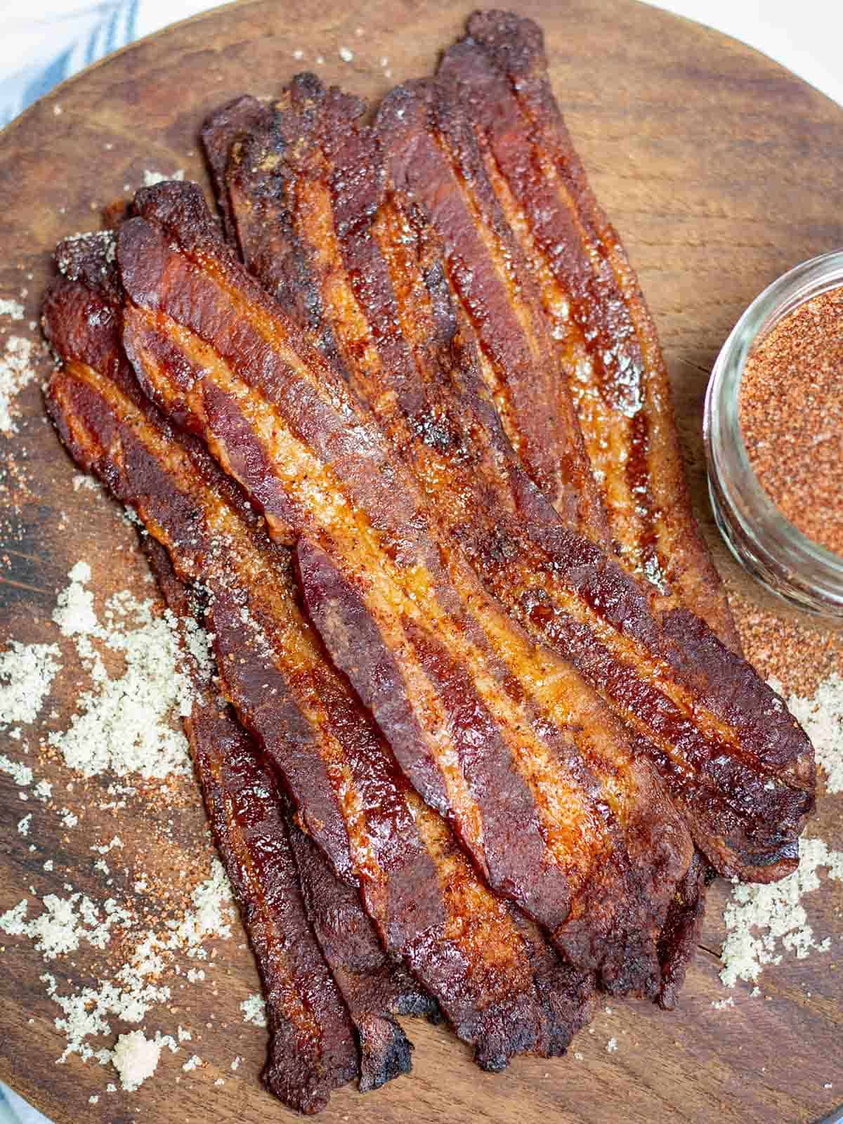 Candied bacon on a brown board with sprinkling of brown sugar and a jar of dry rub nearby.